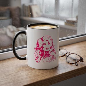 Puede incluir: Una taza de cerámica blanca con asa y borde negros, que presenta una ilustración gráfica en rosa intenso de una mujer con cabello voluminoso y la firma Dolly con un pequeño corazón debajo. Unas gafas de carey marrón descansan al lado.
