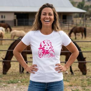 May include: A white short-sleeved t-shirt featuring a hot pink graphic print of a woman with voluminous hair on the chest. The graphic includes the signature Dolly and a small heart symbol below the portrait, centered on the front of the garment.