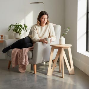 May include: A light-coloured wooden side table with a round top and three angled legs, featuring a smooth finish. The table stands on a grey floor and holds a small white ceramic vase with a single green leafy stem, showcasing a simple design.