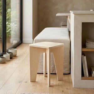 Pode incluir: Um banco de madeira clara com um topo quadrado e quatro pernas anguladas. O banco é construído em contraplacado com padrões de grão visíveis. Apresenta um design simples e funcional com um acabamento liso sobre um piso de madeira clara.