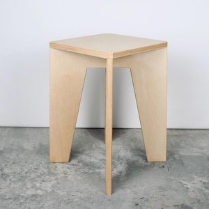 Puede incluir: Un taburete de madera de color claro con un asiento cuadrado y tres patas. Las patas están en ángulo, y el soporte central es una sola pieza vertical. El taburete está hecho de madera contrachapada y tiene un acabado natural.