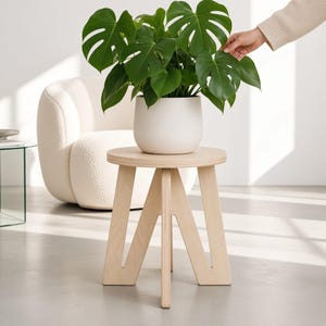 May include: A light-coloured wooden stool with a circular top and three V-shaped legs supporting a white ceramic planter containing a green Monstera plant with large, split leaves. The stool features a smooth, natural wood grain finish.