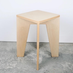May include: A light-coloured wooden stool with a triangular top and three legs. The legs are angled outwards, and the central leg is straight. The stool is made of layered wood and has a simple, modern design.