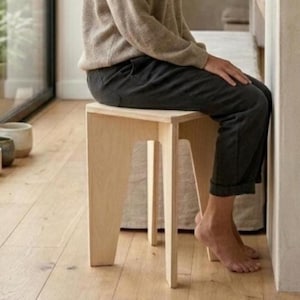 May include: A light-colored wooden stool with four angled legs and a flat square seat. The stool is constructed from light plywood with visible grain patterns, featuring a simple geometric design with open sides and a sturdy base structure.