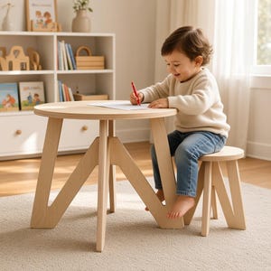 Puede incluir: Una mesa redonda de madera clara con patas inclinadas y un taburete pequeño a juego. La mesa cuenta con una superficie circular lisa, sostenida por tres patas de madera distintivas, anchas y en forma de V que proporcionan una base estable.