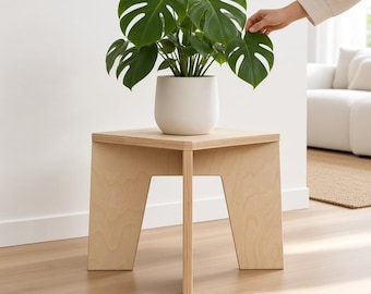 May include: A light-colored wooden side table with a square top and three angled legs. A white ceramic pot containing a green Monstera plant with large, split leaves sits on the table surface. The table features visible plywood grain edges.