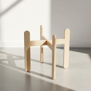 May include: A light-colored wooden plant stand featuring a cross-shaped base design with four vertical legs and rounded top edges. The structure is constructed from interlocking flat wooden panels, providing a stable frame for holding items.