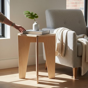 May include: A light-colored wooden side table with a triangular top and three angled legs. A small white ceramic vase containing a single green monstera leaf and a thick book with a dark cover are placed on the flat surface of the wooden table.