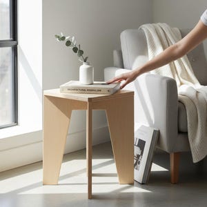 May include: A light-colored wooden side table with three angled legs and a triangular top. A white ceramic vase containing a green eucalyptus sprig and a book titled ARCHITECTURAL MINIMALISM are placed on the surface of the wooden table.