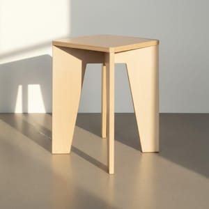 May include: A light-coloured wooden stool with a square top and four angled legs. The stool is constructed from plywood, showing visible layered edges on the seat and legs. The design features a clean, geometric structure with a smooth finish.