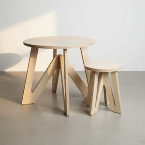 May include: A light-colored wooden round table and a matching round stool. Both pieces feature a smooth, natural wood grain finish and are constructed with angled, V-shaped legs that provide structural support for the circular tops.