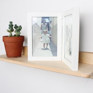 May include: A light-colored wooden shelf with a terracotta pot holding a small succulent plant. A white double picture frame displays a photograph. The shelf is mounted on a white wall.