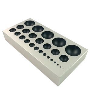 May include: A rectangular, silver-colored metal forming block with a series of graduated, black, hemispherical depressions. The depressions range in size from small to large, arranged in a descending pattern.