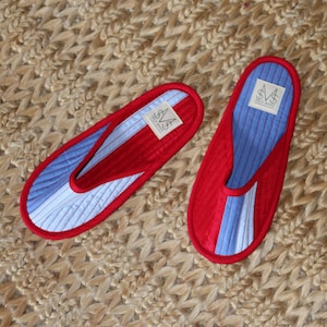 May include: A pair of red slippers with blue and white striped interiors. The slippers have a quilted design and a small label on the inside. They are displayed on a textured, woven surface.