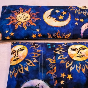 May include: Two blue fabric pouches with a sun and moon design. The pouches are lined up side by side. The sun and moon are yellow, orange, and white. The background is a dark blue with white stars.