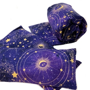 May include: Three purple and gold fabric eye pillows with a celestial design. The pillows feature a constellation pattern with stars, planets, and a crescent moon.