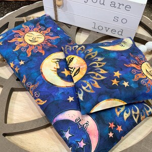 May include: Two blue fabric heat packs with a sun and moon design. The fabric has a watercolor look with stars and a blue background. The text "You are so loved" is visible in the background.
