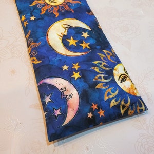 May include: A blue fabric pouch with a repeating pattern of suns and moons. The suns are yellow with orange rays and the moons are white with a yellow crescent shape. The background is a dark blue with white stars.