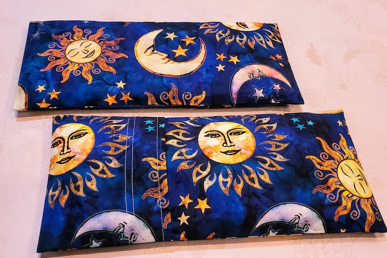 May include: Two blue fabric pouches with a sun and moon design. The pouches are lined up side by side. The sun and moon are yellow, orange, and white. The background is a dark blue with white stars.