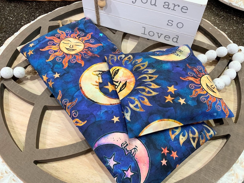 May include: Two blue fabric heat packs with a sun and moon design. The fabric has a watercolor look with stars and a blue background. The text "You are so loved" is visible in the background.