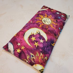 May include: A purple and gold fabric eye pillow with a celestial pattern featuring moons, stars, and a sun with a face.
