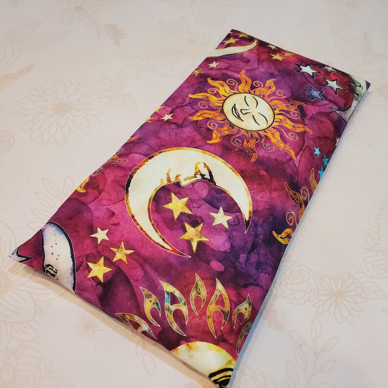 May include: A purple and gold fabric eye pillow with a celestial pattern featuring moons, stars, and a sun with a face.