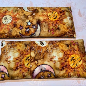 May include: Two brown fabric heat packs with a sun and moon design. The fabric has a watercolor look with gold, pink, and white accents. The sun and moon have faces.