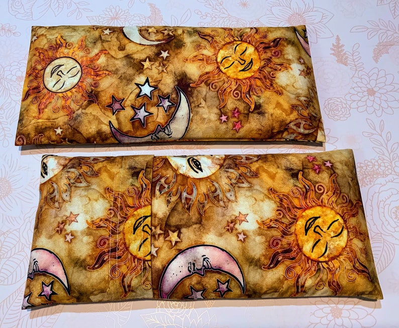 May include: Two brown fabric heat packs with a sun and moon design. The fabric has a watercolor look with gold, pink, and white accents. The sun and moon have faces.