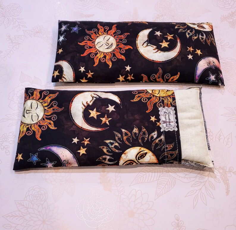 May include: Two black fabric eye pillows with a white background and a pattern of suns and moons. The pillows are filled with flax seeds and lavender. The pillows are handmade and are perfect for relaxation and sleep.