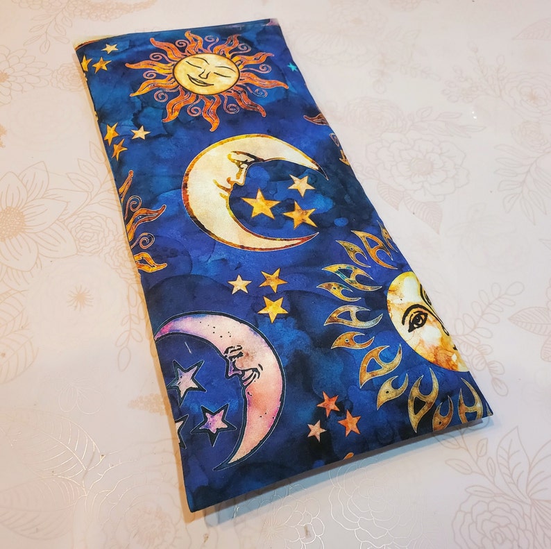 May include: A blue fabric pouch with a repeating pattern of suns and moons. The suns are yellow with orange rays and the moons are white with a yellow crescent shape. The background is a dark blue with white stars.