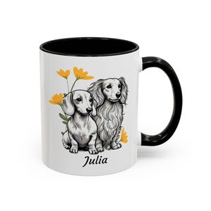 Può includere: Tazza in ceramica bianca con interno e manico neri. La tazza presenta un'illustrazione in bianco e nero di due bassotti con fiori gialli. Il nome "Julia" è stampato sotto i cani.