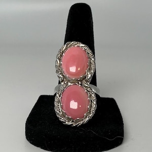 May include: A silver ring featuring two vertically stacked, oval-shaped pink cabochon stones. Each stone is set within a twisted rope-style metal border, creating a continuous elongated design that sits prominently on the finger band.