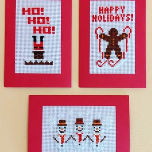 Könnte beinhalten: Drei handgefertigte Weihnachtskarten mit roten Rändern. Die obere linke Karte zeigt "HO! HO! HO!" mit einem Weihnachtsmann-Design. Die obere rechte Karte zeigt "FRÖHLICHE WEIHNACHTEN!" mit einem Lebkuchenmann. Die untere Karte zeigt drei Schneemänner.