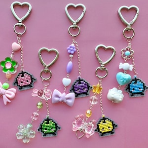 May include: Five keychains with heart-shaped clasps and decorative charms. Each keychain features a pixelated character, with charms including bows, flowers, hearts, and beads in various colors like pink, purple, and blue. The background is a solid pink.