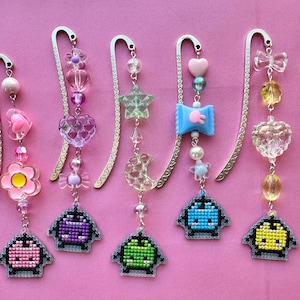 May include: Five decorative metal bookmarks with colorful beaded charms and pixelated alien designs. Charms include flowers, hearts, stars, bows, and moons in pink, purple, blue, green, and yellow. The bookmarks are on a pink background.
