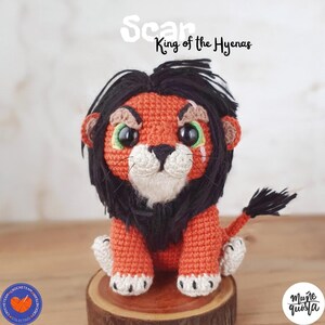 K&ouml;nnte beinhalten: Geh&auml;kelte L&ouml;wenpuppe in Orange, Schwarz und Wei&szlig; mit gr&uuml;nen Augen. &Uuml;ber der Puppe steht der Text "Scar King of the Hyenas". Die Puppe sitzt auf einem Holzsockel. Die Puppe ist handgefertigt.
