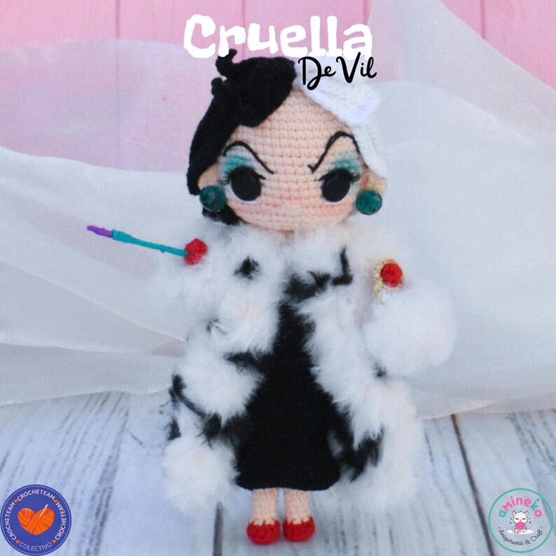 K&ouml;nnte beinhalten: Geh&auml;kelte Puppe von Cruella De Vil, mit schwarz-wei&szlig;er Frisur, gr&uuml;nen Ohrringen und einem schwarzen Kleid mit wei&szlig;em Pelzmantel. Die Puppe h&auml;lt ein kleines, farbenfrohes Accessoire. Der Text "Cruella De Vil" steht dar&uuml;ber.