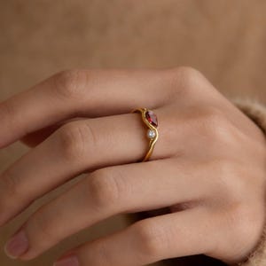 May include: A gold ring featuring a pear-shaped red gemstone and a small, round, clear gemstone. The ring is on a person's finger, with a soft, neutral-toned background.