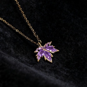 May include: A gold-colored necklace with a pendant shaped like a maple leaf. The leaf is filled with purple gemstones. The necklace is displayed on a black velvet surface.
