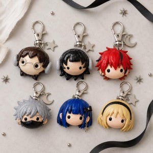 May include: Six chibi-style character keychains featuring sculpted heads with distinct hair colors including brown, black, red, silver, blue, and blonde. Each keychain includes a silver metal lobster clasp and small charms like stars or moons.