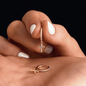 Puede incluir: Pendientes de aro dorados con colgantes de cruz. Los aros y las cruces están incrustados con pequeñas piedras brillantes. Los pendientes se muestran sobre un fondo oscuro.