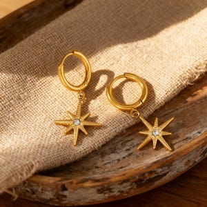 May include: Gold-colored hoop earrings with a starburst pendant. Each earring features a small, clear gemstone at the center of the star. The earrings are displayed on a tan fabric inside a wooden tray.