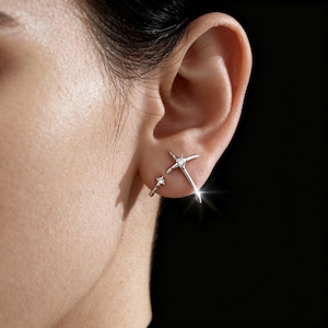 May include: A pair of silver earrings. One earring is a small hoop with a single clear stone. The other earring is a starburst design with a clear stone in the center. The earrings are worn on the ear.