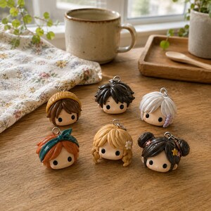 May include: Six small, round, sculpted character head charms with metal loops. The figures feature diverse hair colors including brown, black, white, orange, and blonde, styled with accessories like a yellow beanie, teal headband, and small flowers.