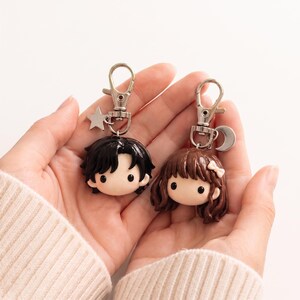 May include: Two character keychains featuring sculpted heads with smooth, pale skin and black eyes. One has short black hair with a silver star charm, and the other has wavy brown hair with a small white bow and a silver crescent moon charm attached.