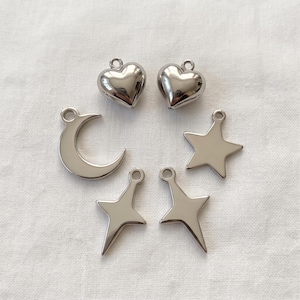 May include: A collection of six silver-toned metal charms featuring two puffed hearts, one crescent moon, one five-pointed star, and two four-pointed star shapes. Each charm includes a small circular loop at the top for attachment purposes.