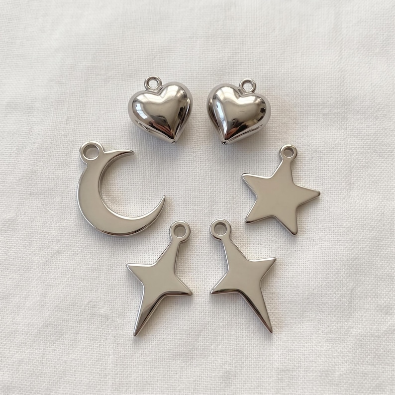 May include: A collection of six silver-toned metal charms featuring two puffed hearts, one crescent moon, one five-pointed star, and two four-pointed star shapes. Each charm includes a small circular loop at the top for attachment purposes.