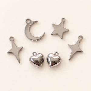 May include: A collection of six silver-toned metal charms featuring a crescent moon, a five-pointed star, two four-pointed star shapes, and two puffed heart shapes. Each charm includes a small circular loop at the top for attachment purposes.