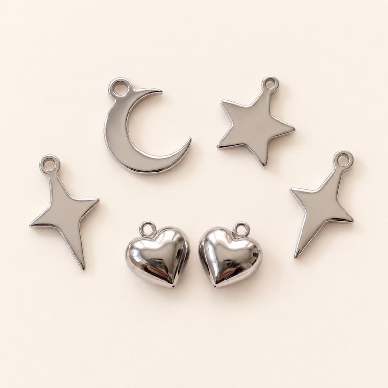 May include: A collection of six silver-toned metal charms featuring a crescent moon, a five-pointed star, two four-pointed star shapes, and two puffed heart shapes. Each charm includes a small circular loop at the top for attachment purposes.