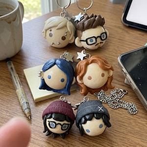 May include: Six miniature character head keychains made of smooth material, each featuring a silver star charm and a metal ring. The heads display various hair colors, styles, and facial features, including glasses, beanies, and piercings, arranged in three rows.